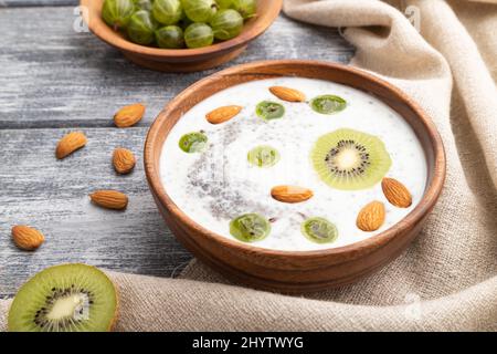 Joghurt mit Kiwi, Stachelbeere, Chia und Mandeln in Holzschüssel auf grauem Holzhintergrund und Leinentextilien. Seitenansicht, Nahaufnahme. Stockfoto