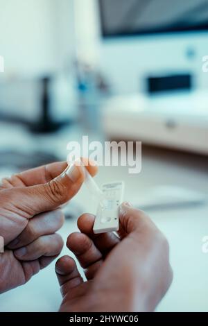 Nahaufnahme eines jungen Mannes, der seine eigene Probe aus einer Pipette in das Schnellantigen-Diagnosetestgerät Covid-19 im Büro legt Stockfoto