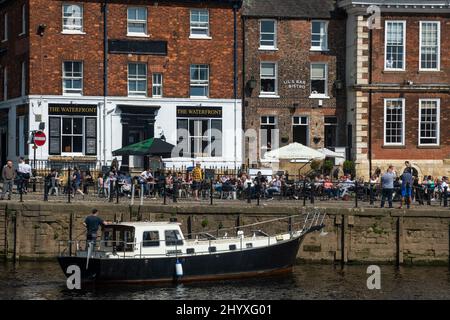 Gäste trinken, essen in belebten Cafés am Flussufer und segeln auf einem Freizeitboot - der malerische sonnige River Ouse, King's Staith, York, North Yorkshire, England. Stockfoto