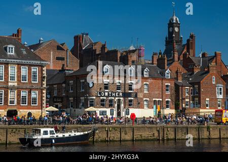 Gäste trinken, essen im Freien in der Sonne in einem geschäftigen Pub am Flussufer und ein Freizeitschiff, das vor Anker liegt - River Ouse, King's Staith, York, North Yorkshire, England. Stockfoto