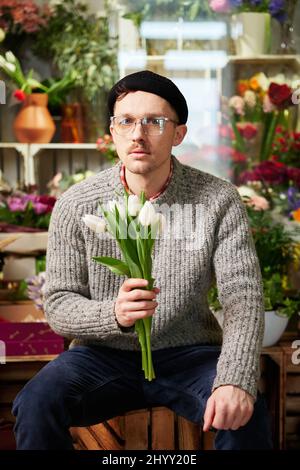 Attraktiver junger Kaukasier mit Brillen und Beanie im Blumengeschäft mit Tulpenstrauß. Gut aussehende männliche Floristin in Pullover hält Strauß. Konzept für kleine Unternehmen. Hochwertige Fotos Stockfoto