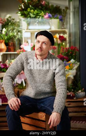 Erfolgreiche stilvolle Unternehmer kaukasischen männlichen Floristen trägt Pullover, Beanie und Brille im Blumenladen. Junger hübscher Gewächshausbesitzer oder Gärtner, der Blumen und Blumensträuße im Geschäft verkauft Stockfoto