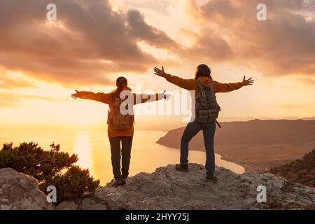 Zwei junge Wanderer stehen mit erhobenen Armen auf dem Gipfel des Berges und blicken auf das Meer und den Sonnenuntergang Stockfoto