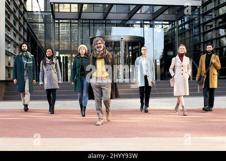 Eine multiethnische Gruppe von fröhlichen Geschäftsleuten, die vor einem Unternehmenszentrum auf die Kamera zulaufen. Stockfoto