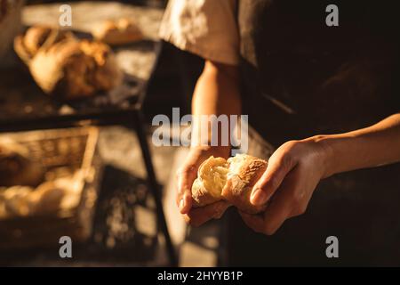 Mittlerer Abschnitt der asiatischen mittleren erwachsenen weiblichen Bäckerin, die im Stand in der Bäckerei Brot bricht Stockfoto