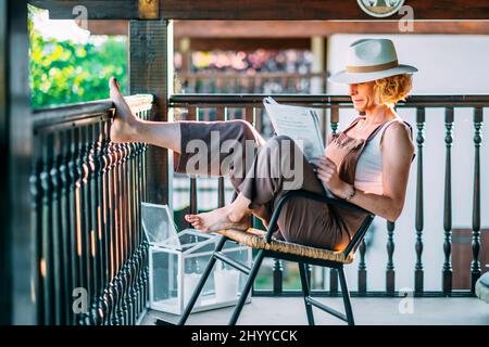Porträt einer reifen kaukasischen blonden jungen Frau im Alter von 50s Jahren, die einen Hut trägt und eine Zeitung liest und in entspannter Pose auf einem Schaukelstuhl sitzt Stockfoto
