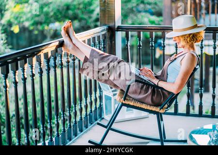 Porträt einer reifen kaukasischen blonden jungen Frau im Alter von 50s Jahren mit einem Laptop, der einen Hut trägt und in entspannter Pose in einem Schaukelstuhl auf dem Holzboden sitzt Stockfoto