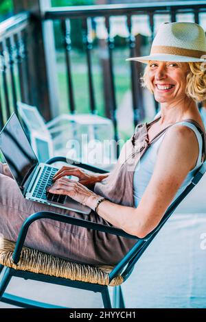 Porträt einer reifen kaukasischen blonden jungen Frau im Alter von 50s Jahren mit einem Laptop, der einen Hut trägt und in entspannter Pose in einem Schaukelstuhl auf dem Holzboden sitzt Stockfoto