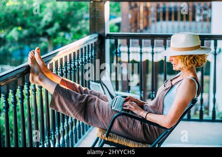 Porträt einer reifen kaukasischen blonden jungen Frau im Alter von 50s Jahren mit einem Laptop, der einen Hut trägt und in entspannter Pose in einem Schaukelstuhl auf dem Holzboden sitzt Stockfoto