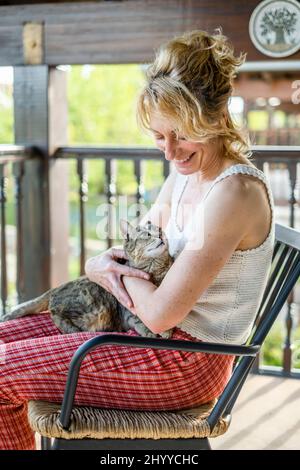 Porträt einer jungen reifen kaukasischen blonden Frau, die mit ihrer Katze zu Hause auf der Veranda lächelt und in einem Schaukelstuhl sitzt. Lifestyle-Konzept. Stockfoto