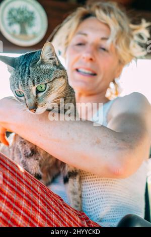 Porträt einer jungen reifen kaukasischen blonden Frau, die mit ihrer Katze zu Hause auf der Veranda lächelt und in einem Schaukelstuhl sitzt. Lifestyle-Konzept. Stockfoto