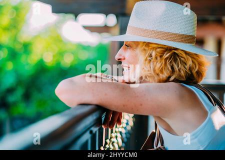 Porträt einer reifen kaukasischen blonden jungen Frau in den Fünfzigern, die einen Hut auf der hölzernen Veranda eines Landhauses trägt. Lifestyle-Konzept. Stockfoto