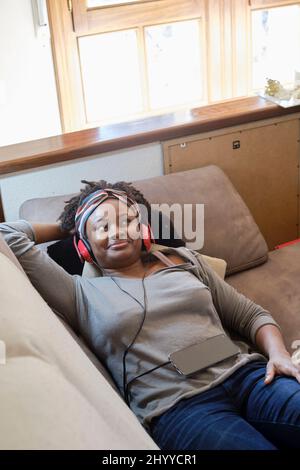 Porträt einer jungen schwarzen Frau, die mit Kopfhörern im Innenbereich Musik hört und auf einem Sofa liegt. Lifestyle-Konzept. Stockfoto