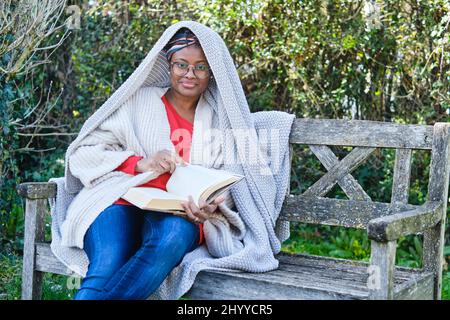 Porträt einer jungen schwarzen Frau mit Afro-Frisur und Brille beim Lesen eines Buches, das auf einer alten Gartenbank sitzt. Lifestyle-Konzept. Stockfoto