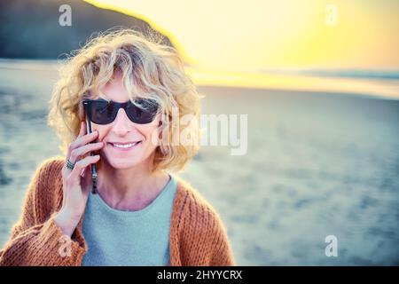 Junge reife blonde kaukasische Frau, die mit einem Mobiltelefon im Freien an einem Strand spricht. Berria Beach, Kantabrien, Spanien, Europa. Stockfoto