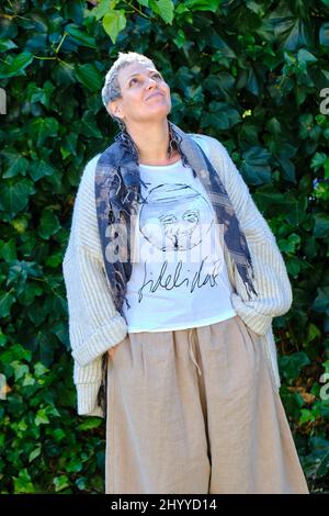 Reife hübsche kaukasische Frau mit grauen kurzen Haaren und lässiger Kleidung, die im Freien in einem Garten mit einem Hintergrund aus Blättern posiert. Stockfoto