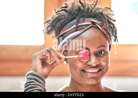 Nahaufnahme Lustiges Porträt einer jungen schwarzen Frau mit Afro-Frisur, die zu Hause mit einem Lutscher in der Halle scherzte und lachte. Lifestyle-Konzept. Stockfoto