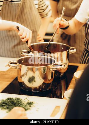 Ungewisse Frauen, die in der Küche zu Hause essen. Eine Frau rührt Suppe in einem Topf auf dem Herd mit einem Löffel. Stockfoto