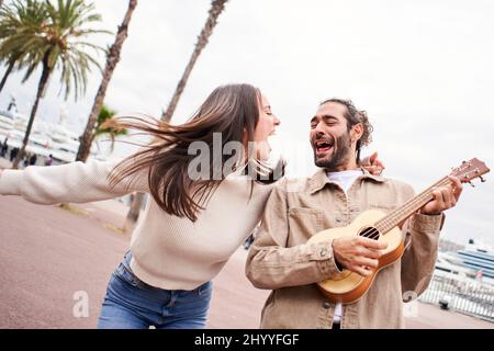 Lustige fröhliche und liebevolle Paar tanzen draußen auf der Straße singen und Gitarre spielen. Mann und Frau haben Spaß während ihres Urlaubs in der Stadt Stockfoto