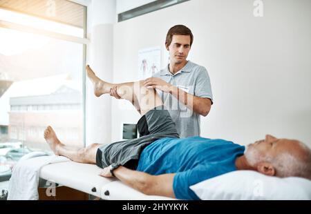 Die Gesundheit des Kunden ist sein einziges Anliegen. Aufnahme eines jungen männlichen Physiotherapeuten, der einem Klienten mit Beinübungen hilft, die auf einem Bett liegen. Stockfoto