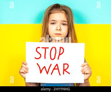 Ein Mädchen weint und hält ein STOP-WAR-Plakat in den Händen vor dem Hintergrund der ukrainischen Flagge. Beendet den Krieg in der Ukraine. Das Konzept des Friedens Stockfoto