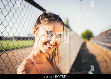 Lächelnde kaukasische junge aktive Frau, die auf die Kamera schaut und eine Musik-Wiedergabeliste auf ihrem Smartphone auflegt Stockfoto