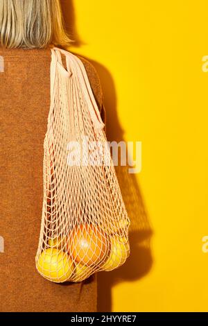 Lebendige Nahaufnahme einer nicht erkennbaren Frau, die Orangen in einer Öko-Einkaufstasche vor gelbem Hintergrund, bewusstem Konsum und Bio-Lebensmittel-Koncep trägt Stockfoto