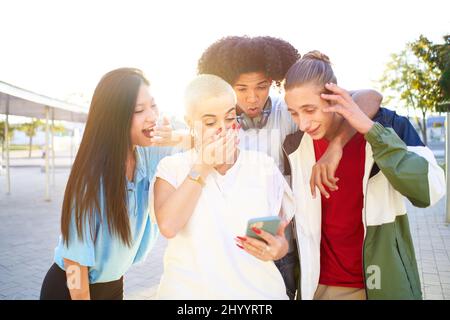 Freunde staunten, als sie das Smartphone im Freien sahen. Junge lächelnde Menschen teilen sich mit mobilen Zellen im Freien. Stockfoto