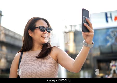 Asiatische Frau in Sonnenbrille nimmt ein Selfie mit dem Handy in einem Einkaufszentrum Stockfoto