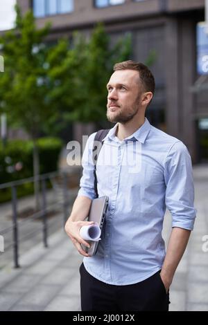 Junger, stilvoller, ernsthafter bärtiger Geschäftsmann, der auf einer Straße mit Laptop und Blaupausen steht. Porträt eines erfolgreichen und selbstbewussten Geschäftsmannes, Designers oder Architekturexperten. Hochwertige Bilder Stockfoto