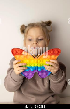 Ein 5-jähriges Mädchen spielt mit einem Pop IT-Spielzeug in Form eines Schmetterlings. Beliebtes Anti-Stress-Spielzeug, das Feinmotorik entwickelt. Konzentrieren Sie sich auf Spielzeug. Stockfoto