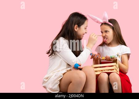 Bild von zwei Schwestern, die sich auf die Osterparty vorbereiten, die Hasenohren tragen und die Nase bemalen, um gemeinsam Spaß zu machen. Stockfoto