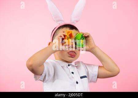 Nahaufnahme eines kleinen niedlichen Jungen mit Hasenohren, der seine Zunge zeigt und seine Augen mit Spielzeug-Eiern auf rosa Hintergrund bedeckt. Stockfoto