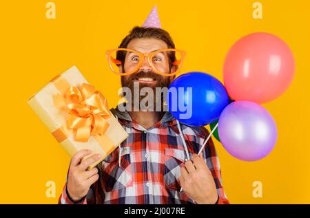 Alles gute zum Geburtstag. Bärtiger Mann in Urlaubsmütze mit Ballons und Geschenk-Box. Stockfoto