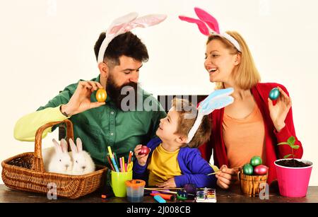 Fröhliche Familie sitzen am Holztisch mit Ostereiern. Familie in Hasenohren bereitet sich auf den Urlaub vor. Stockfoto