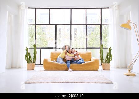 Drei junge Frauen, die nur aus mehreren Rassen kommen, umarmen sich, sitzen im Wohnzimmer auf dem Sofa. Glückliche Dame Mixed Rennen lächelnde Freunde zusammen zu Hause. Stockfoto