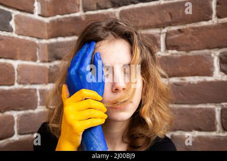 Depressive Frau mit geschlossenen Augen, in der Nähe ihres Gesichts hält die Hände, gemalt in den gelb-blauen Farben der ukrainischen Flagge. Krieg in der Ukraine. Unterstützung für die Ukraine Stockfoto