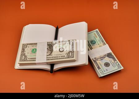 Notebook mit zwei Dollarpaketen auf einem orangefarbenen Tisch. Einsparungskonzept. Fotografieren Sie im horizontalen Format. Speicherplatz kopieren. Stockfoto
