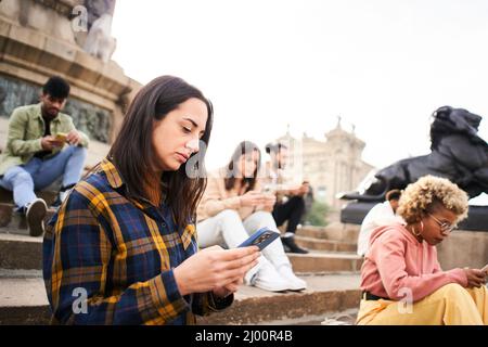 Gruppe von individualistischen Menschen, die im Freien telefonieren und ernsthaftes Gesicht haben. Freunde konzentrierten sich auf ihre Handys, die auf einer Treppe in einer Stadt saßen. Technologie Stockfoto