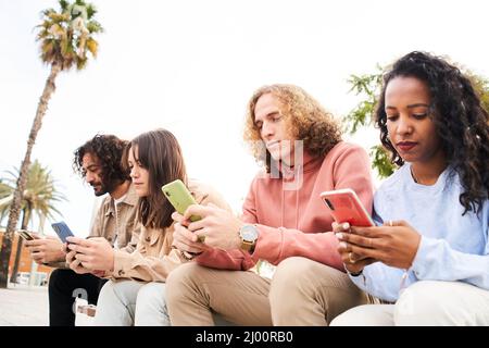 Gruppe von Menschen, die ein Telefon mit ernsthafteren Gesichtes benutzen. Freunde konzentrierten sich auf ihre Handys, die auf einer Bank im Freien in einem Park sitzen. Technologieabhängiges Konzept. Stockfoto