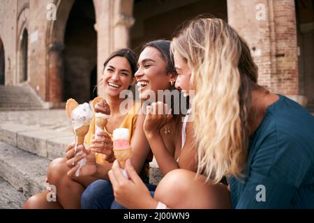 Drei fröhliche junge Freundinnen auf einer Promenade, die fröhlich lächeln, während sie im Sommerurlaub Eiszapfen zum Mitnehmen genießen Stockfoto