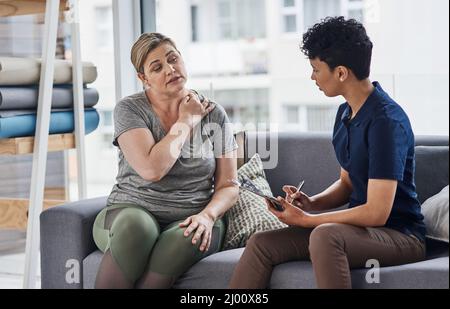 Wann hast du angefangen, diesen Schmerz und dieses Unbehagen zu spüren. Aufnahme einer jungen Physiotherapeutin, die sich mit einer reifen Frau in ihrem Büro in einer Klinik berät. Stockfoto
