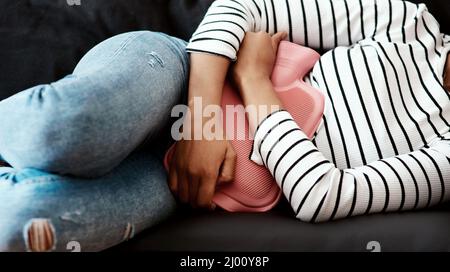 Wenn dein Bauch zum Schlachtfeld wird. Kurzer Schuss einer Frau, die zu Hause auf dem Sofa eine Wärmflasche gegen ihren Bauch hält. Stockfoto