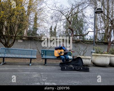 Seattle, WA USA - ca. März 2022: Männlicher Straßenkünstler mit einer akustischen Gitarre, der auf seinem Handy spricht, während er auf einer Bank in der Innenstadt sitzt Stockfoto