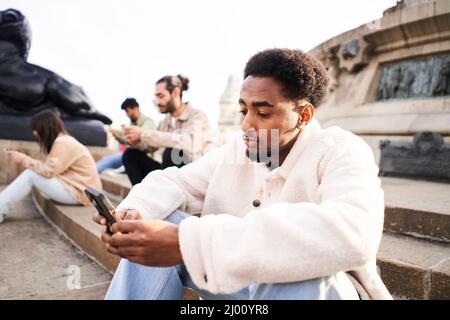 Afroamerikanischer Typ mit mobiler Zelle sitzt in einer Treppe einer Stadt mit anderen Freunden, die sich gegenseitig mit dem Smartphone ignorieren. Technologie-Süchtige Gruppe von Stockfoto