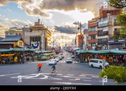 Shijo-dori in der Abenddämmerung mit Radfahrern, Fahrzeugen, Ampeln und einem warmen Leuchten der untergehenden Sonne vom Nishiromon-Tor aus gesehen, Gion, Kyoto, Japan Stockfoto