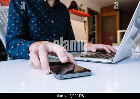Nahaufnahme der Hände eines Mannes mit Laptop, der das Smartphone überprüft Stockfoto