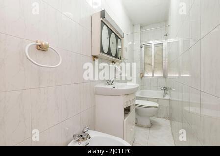Kleines Badezimmer mit weißer Toilettenschüssel, Spiegel am Metallschrank, kleiner Badewanne und Aluminiumfenster und weißen Fliesen Stockfoto