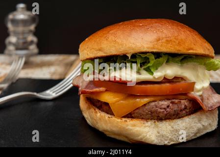 Klassischer Rindfleischburger mit Tomaten, Eisbergsalat, Mayonnaise und Cheddar-Käse auf einem schwarzen Brett mit Gabeln und Salzstreuer auf einem Holztisch Stockfoto