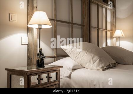 Kopfteil eines Schlafzimmers mit Doppelbetten mit quadratischen Kissen, die mit Holzstangen und einem Nachttisch aus Glas mit Lampe und dekorativem Fuß ausgestattet sind Stockfoto